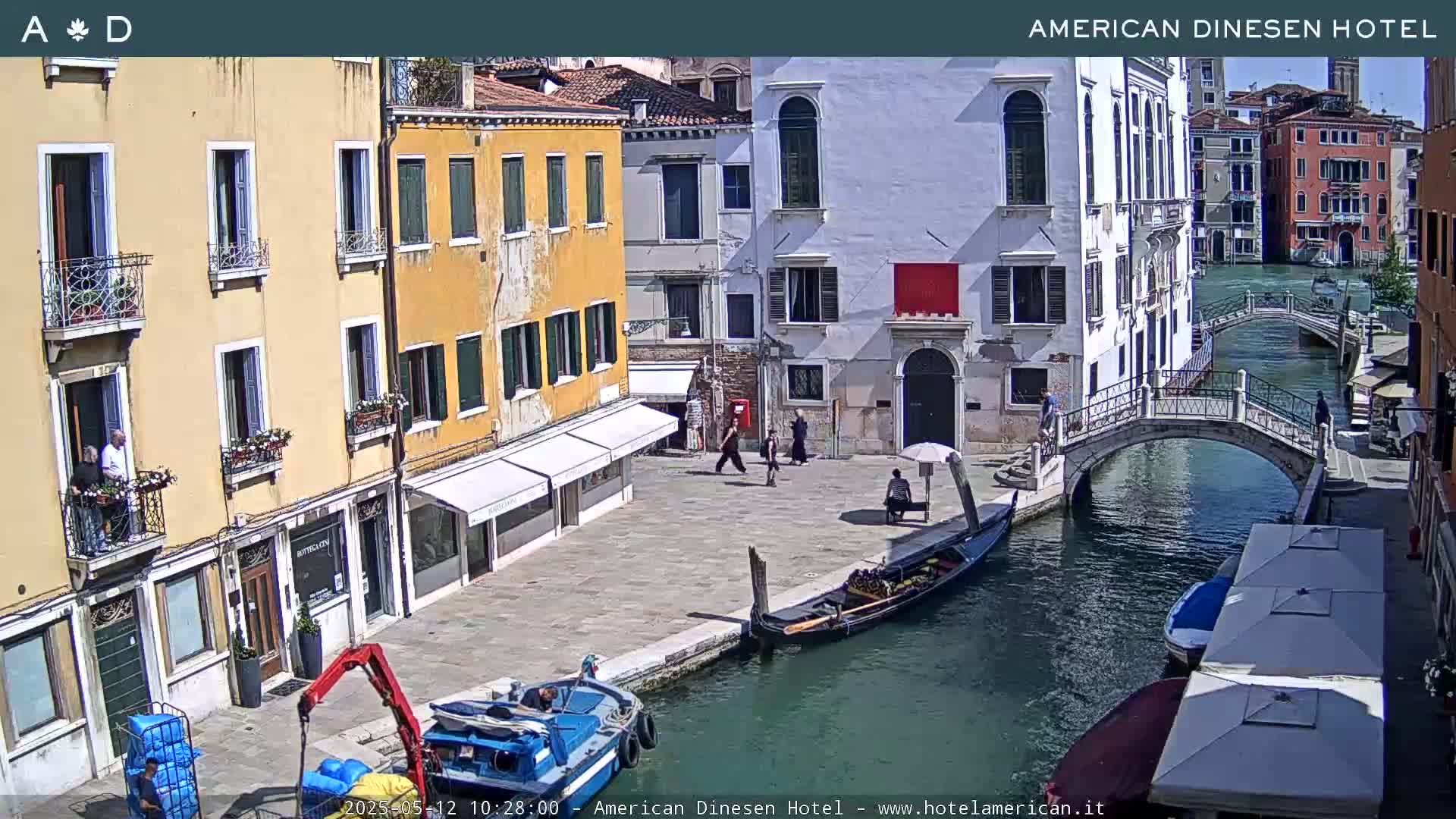 A sunny view of a Venetian canal with gondolas, pedestrians, and colorful buildings lining the waterway, including a small bridge.