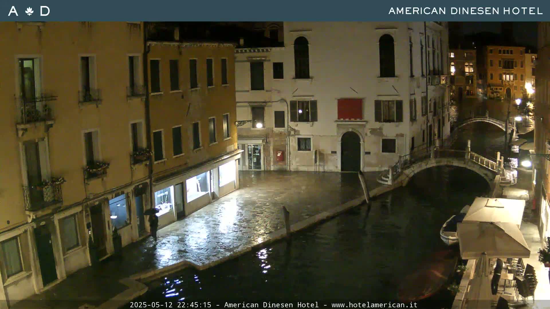 A nighttime view of a canal in Venice, showing several buildings and a bridge, with wet pavement and reflections in the water.
