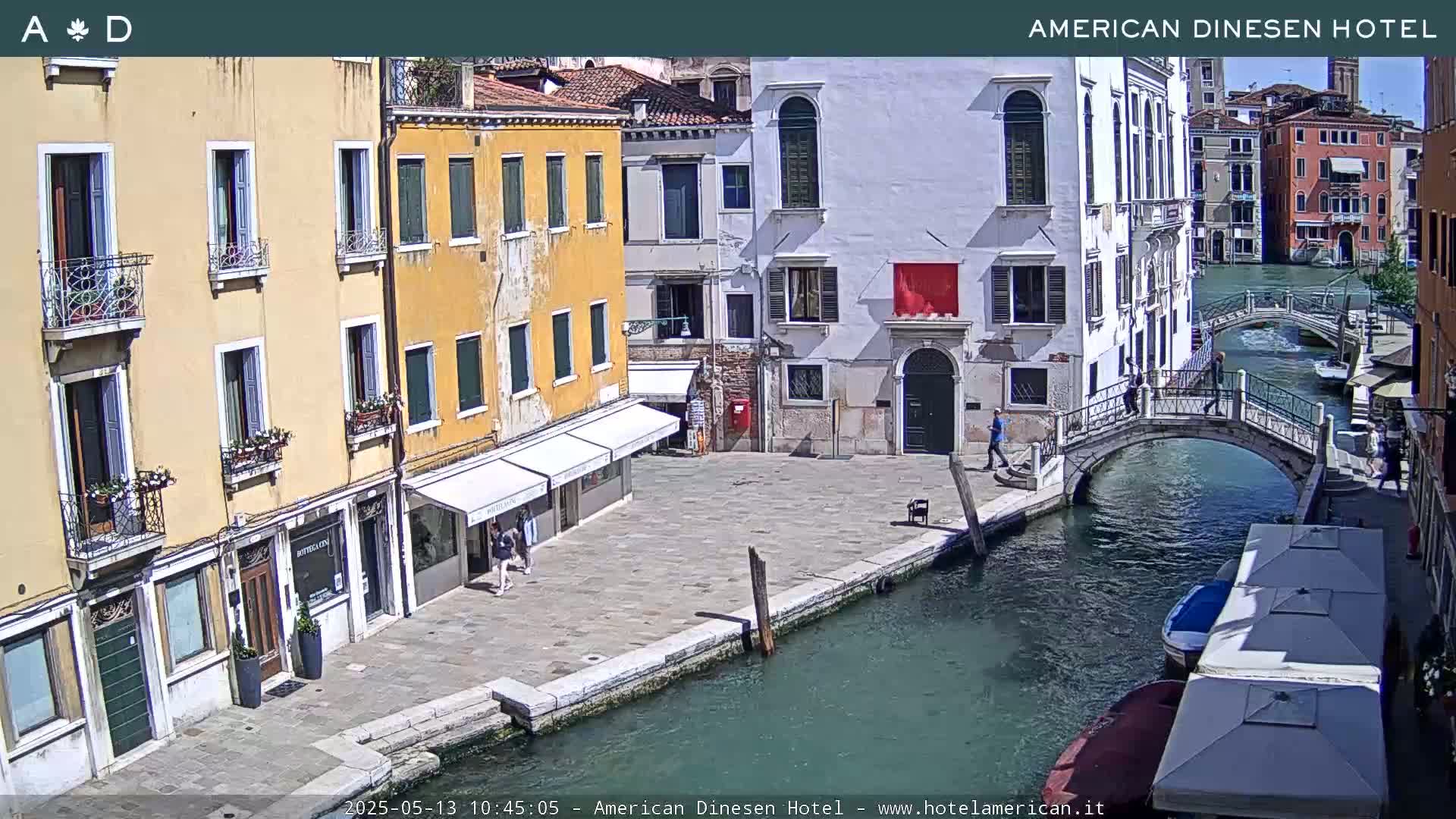 A sunny view of a canal in Venice, Italy, shows several buildings, a footbridge, and a few people walking along the waterfront.