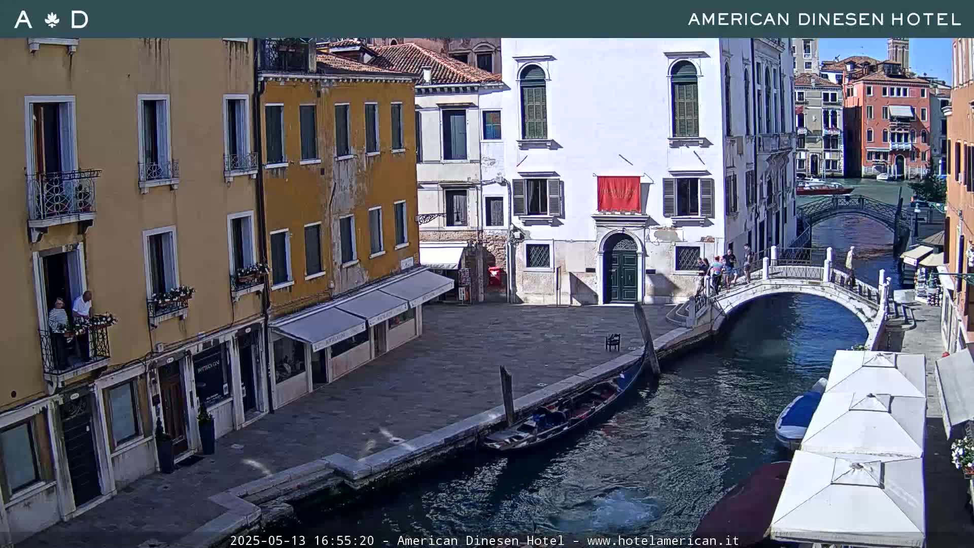 A sunny Venetian canal scene shows colorful buildings lining a waterway, a gondola moored alongside a small bridge with pedestrians, and outdoor cafes.