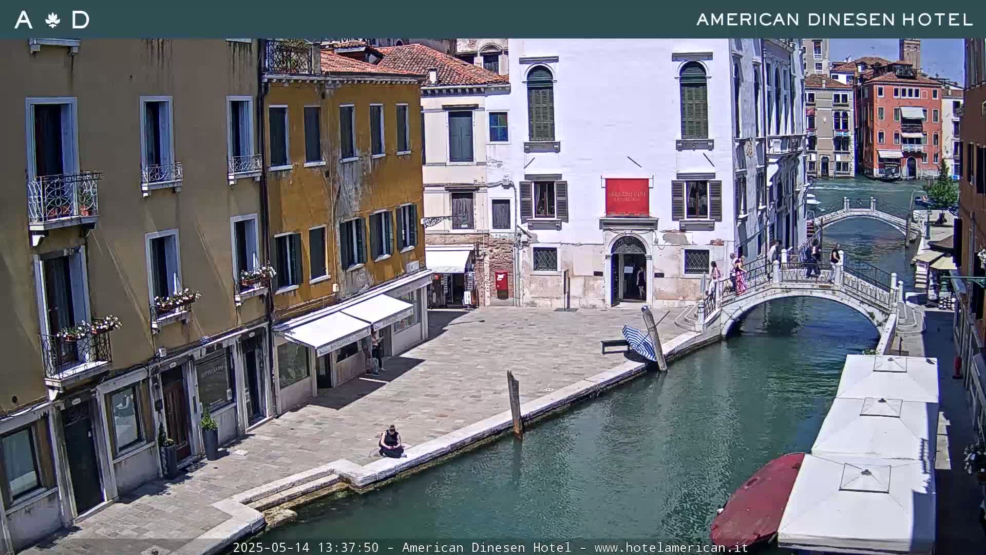 A sunny view of a canal in Venice, Italy, shows colorful buildings lining the waterway, a pedestrian bridge crossing the canal, and several people walking along the shore.