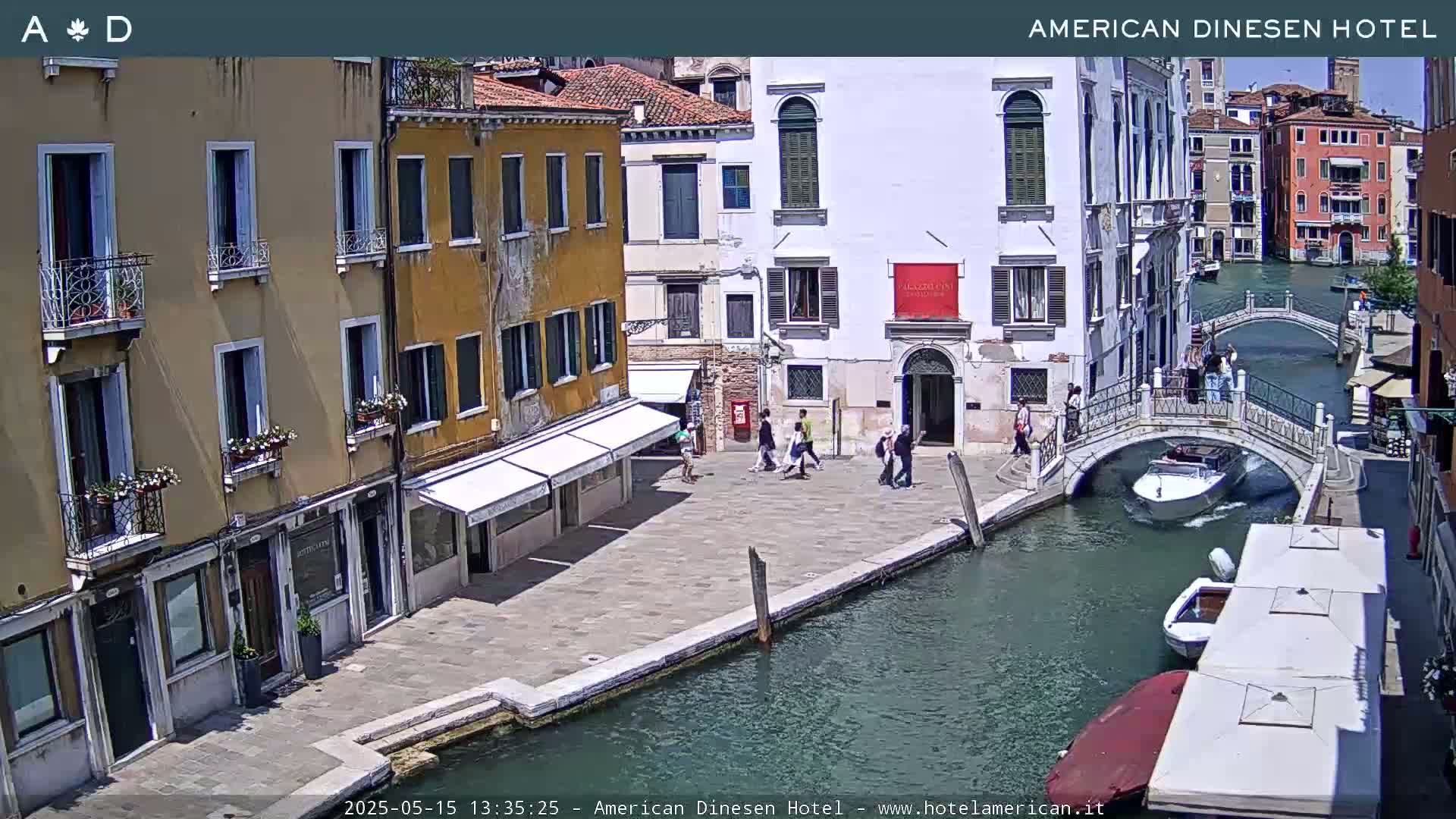 A sunny Venetian canal scene shows several people walking near buildings and a small motorboat on the water, with a pedestrian bridge crossing the canal.