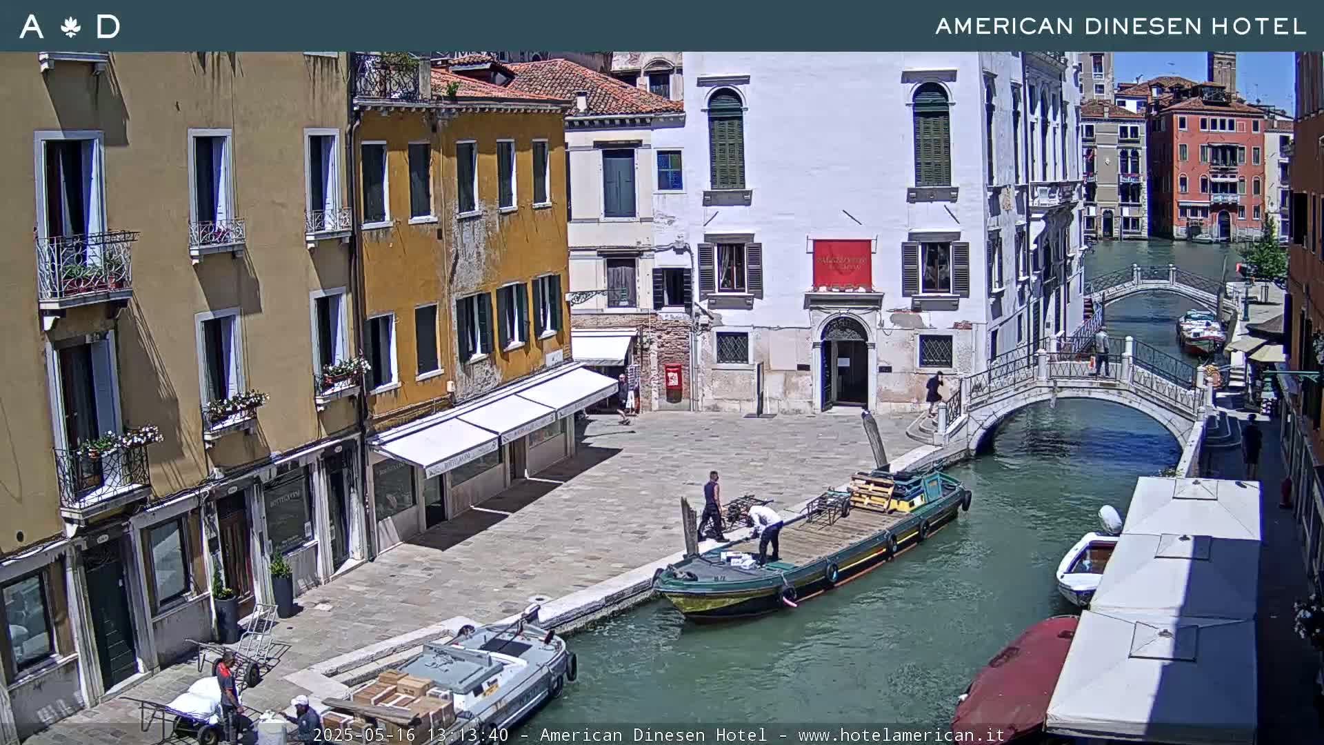 A sunny Venetian canal scene shows several buildings, a small bridge, and a barge being loaded by two people near a waterfront plaza.