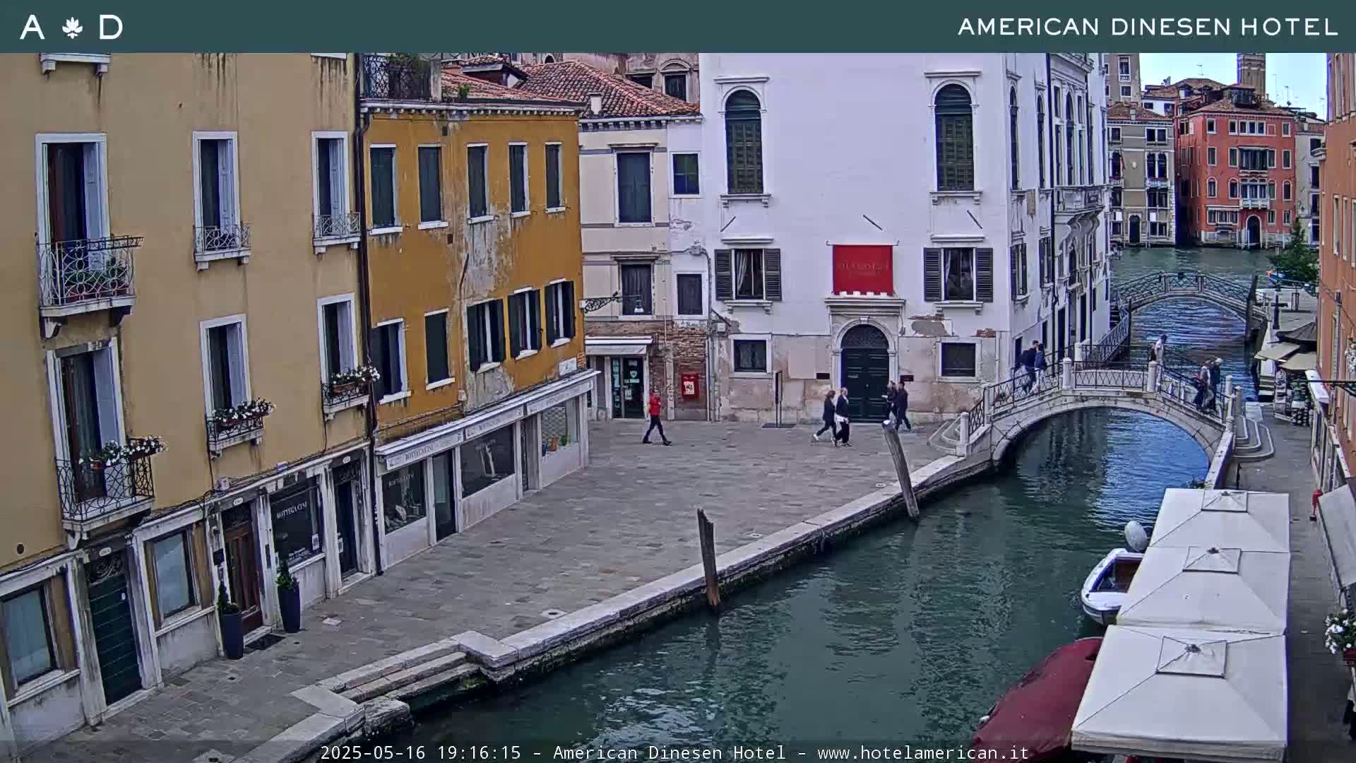 A canal in Venice, under an overcast sky, is lined with buildings and a small footbridge, with a few pedestrians and boats visible.