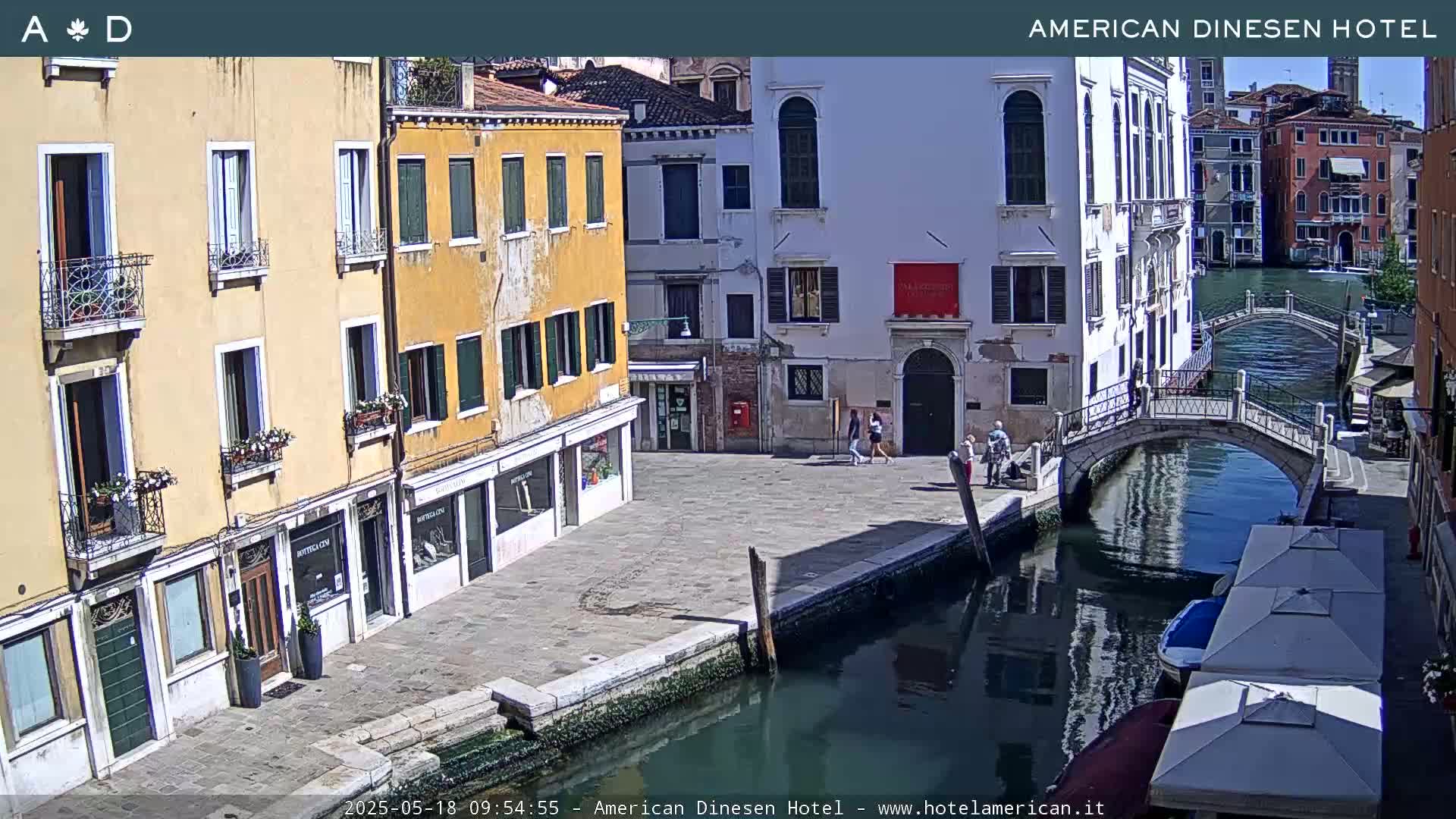 A sunny day in Venice shows a canal with a small bridge, flanked by colorful buildings and a few pedestrians.