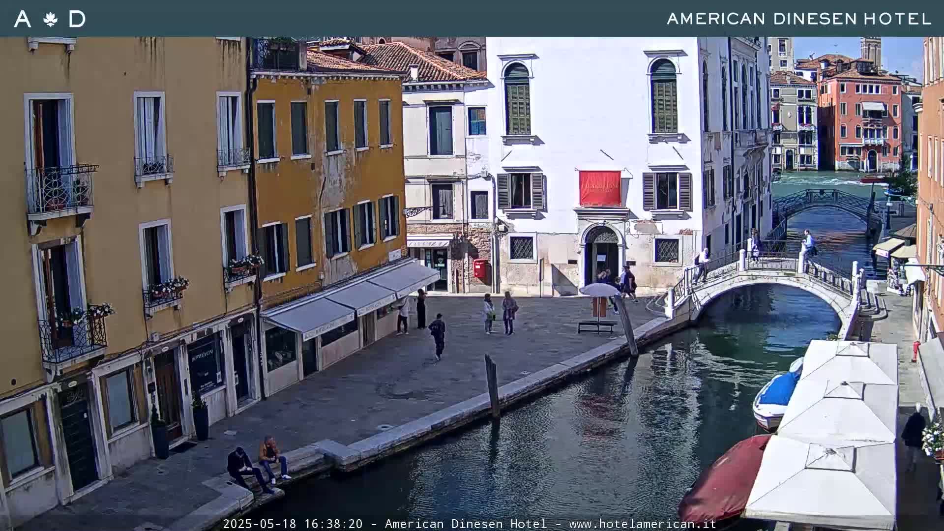 A sunny view of a canal in Venice, Italy, shows a stone bridge, colorful buildings, several people walking and sitting, and boats moored along the canal.