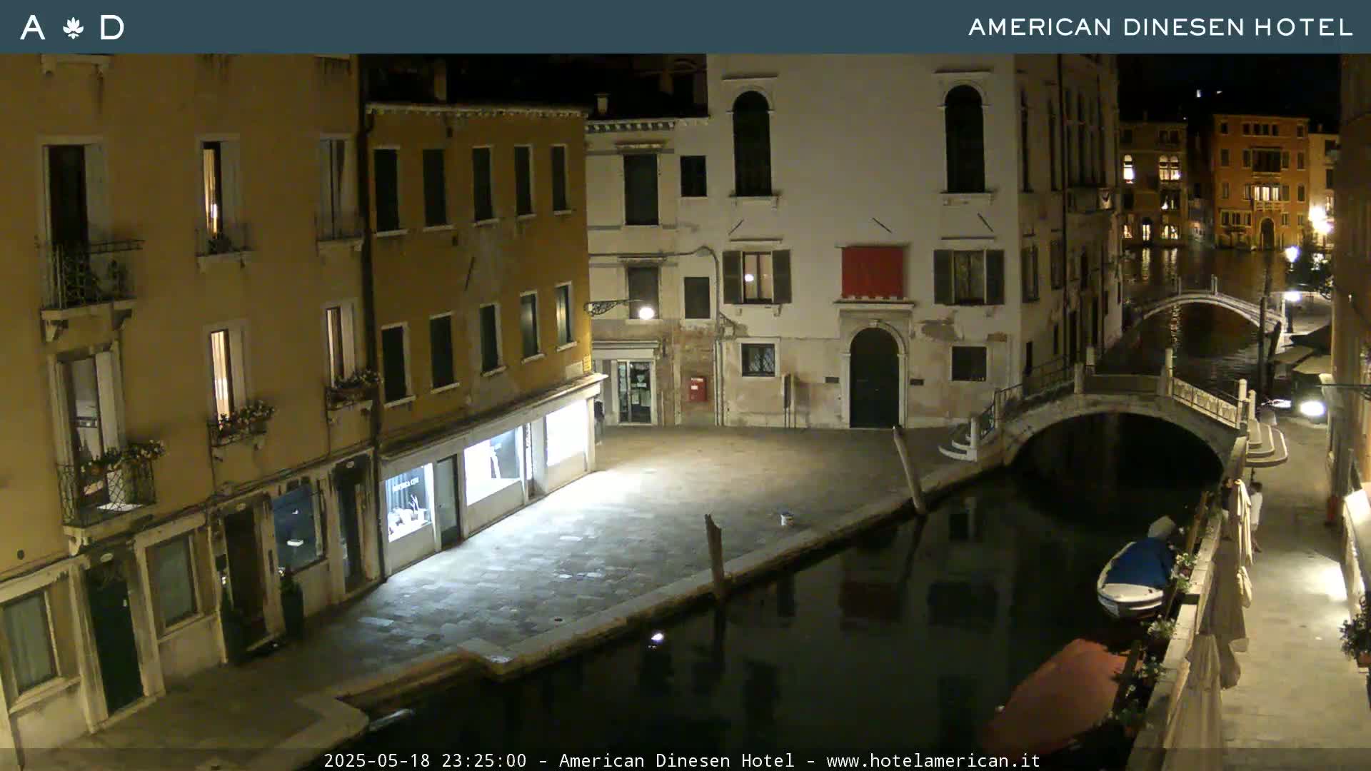 A nighttime view of a canal in Venice, showing several buildings lining the waterway, a small bridge, and a boat moored along the canal's edge.