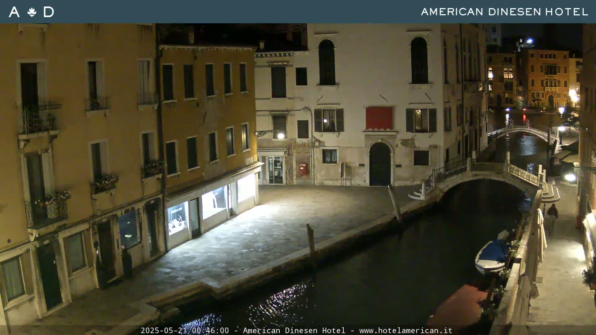 A nighttime view of a canal in Venice, Italy, shows several buildings lining the waterway, with a small bridge and a boat visible on the dark water.