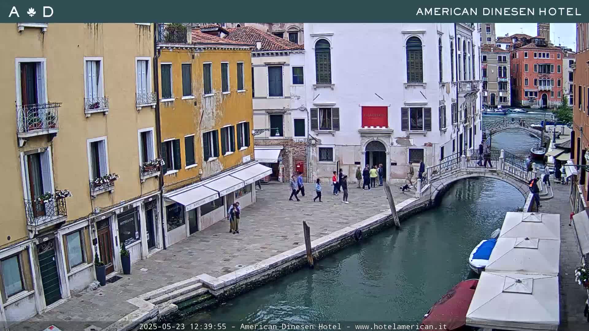 A canal in Venice, Italy, with several people walking along the waterfront and boats on the water, under an overcast sky.