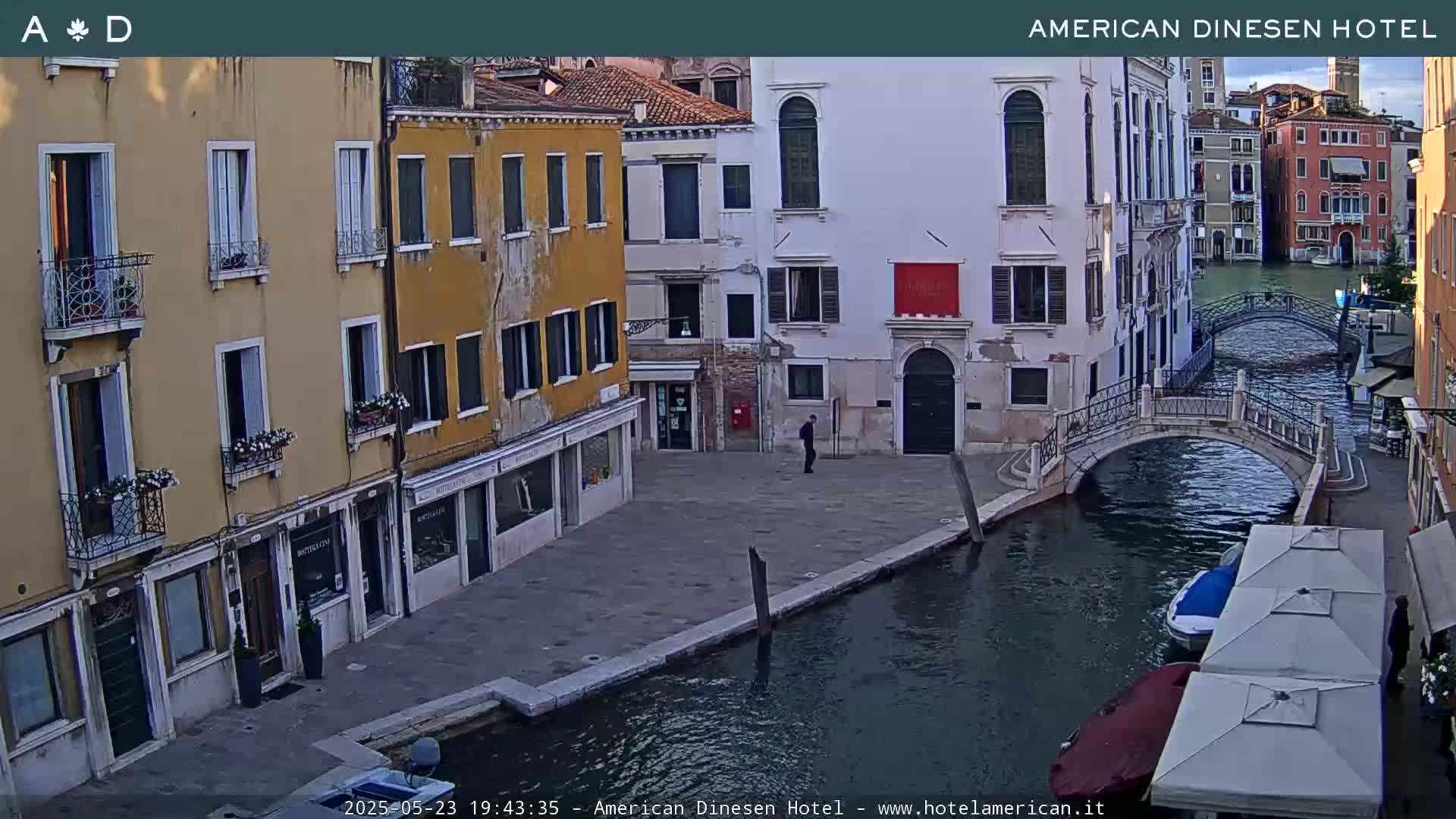 A waterfront view of several buildings in Venice, Italy, with a calm canal, two small bridges, and a person walking on a paved area.