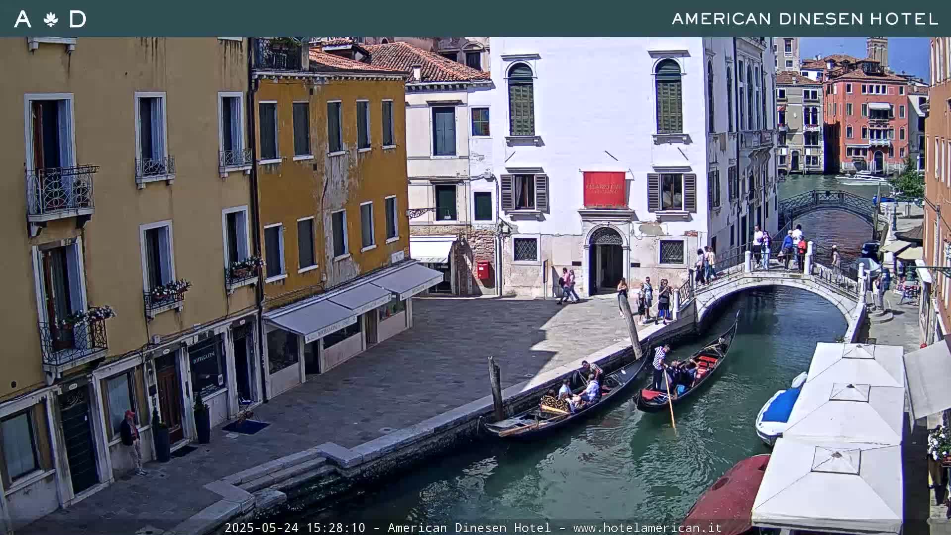 A sunny Venetian canal scene shows gondolas on the water, pedestrians on a bridge and along the canal side, and several buildings lining the waterway.