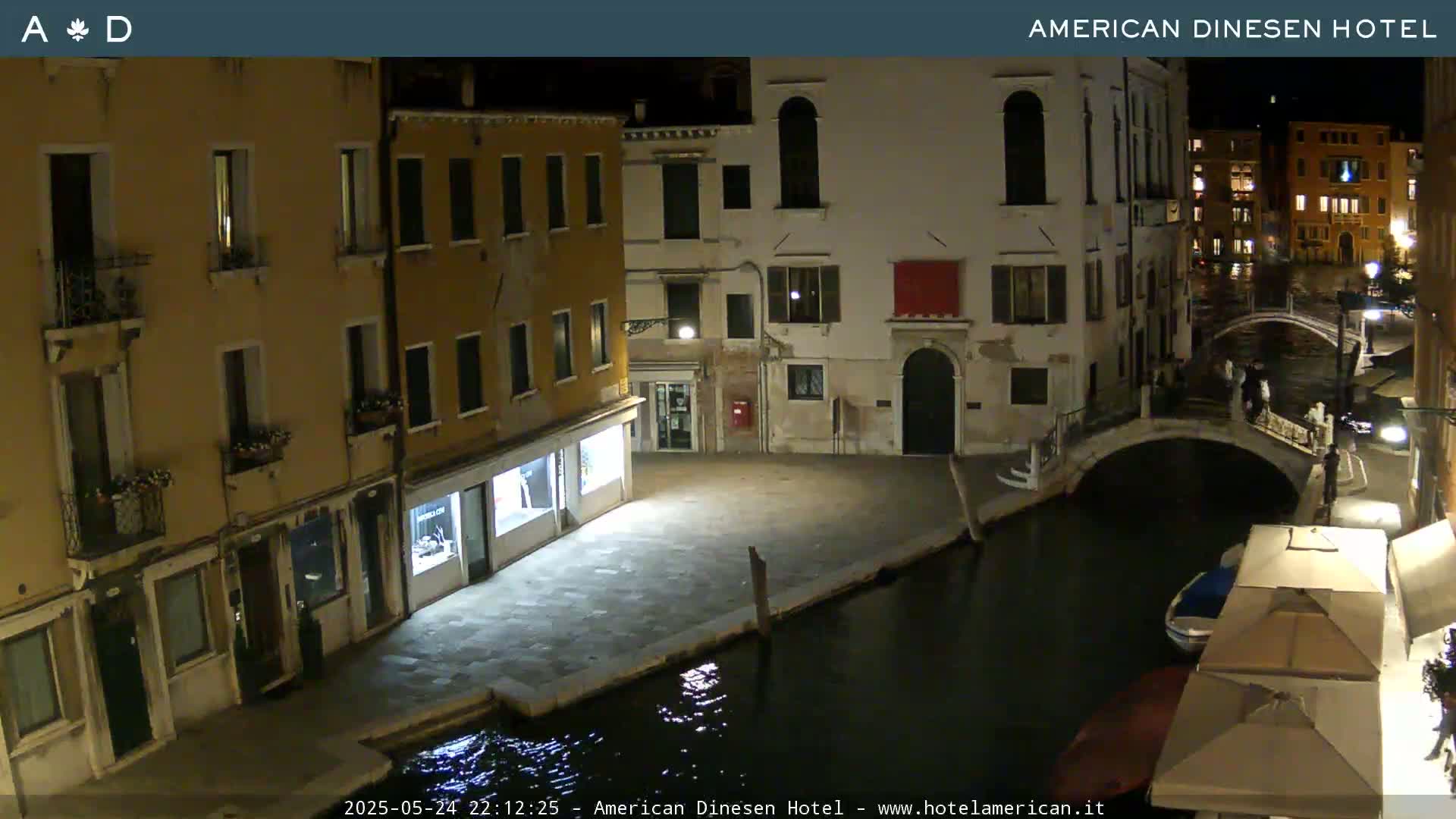 A nighttime view of a canal in Venice, Italy, with several buildings lining the waterway, a small bridge, and a few people visible.