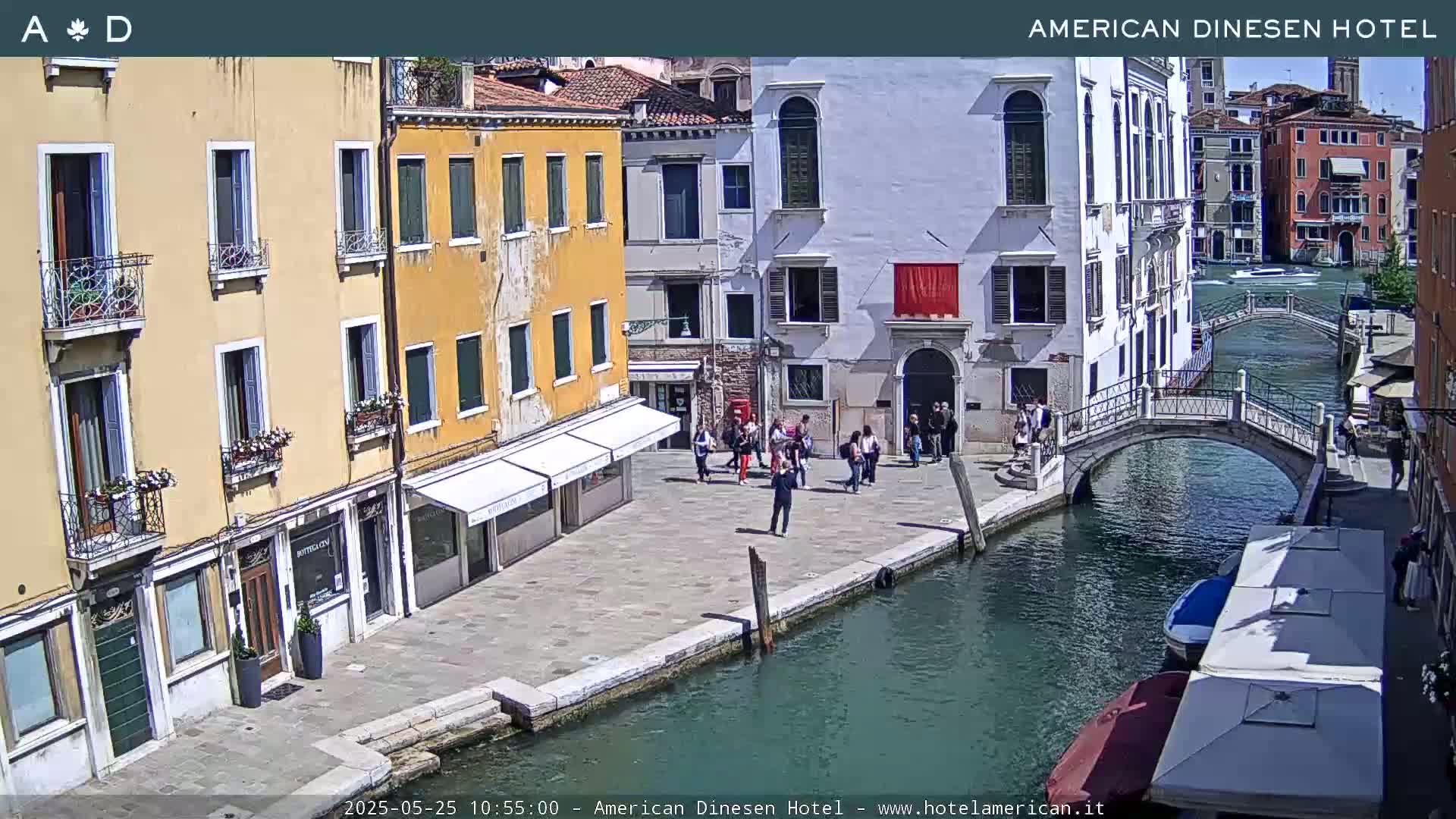 A sunny Venetian canal scene shows several people gathered on a paved walkway beside colorful buildings, with a bridge arching over the waterway.