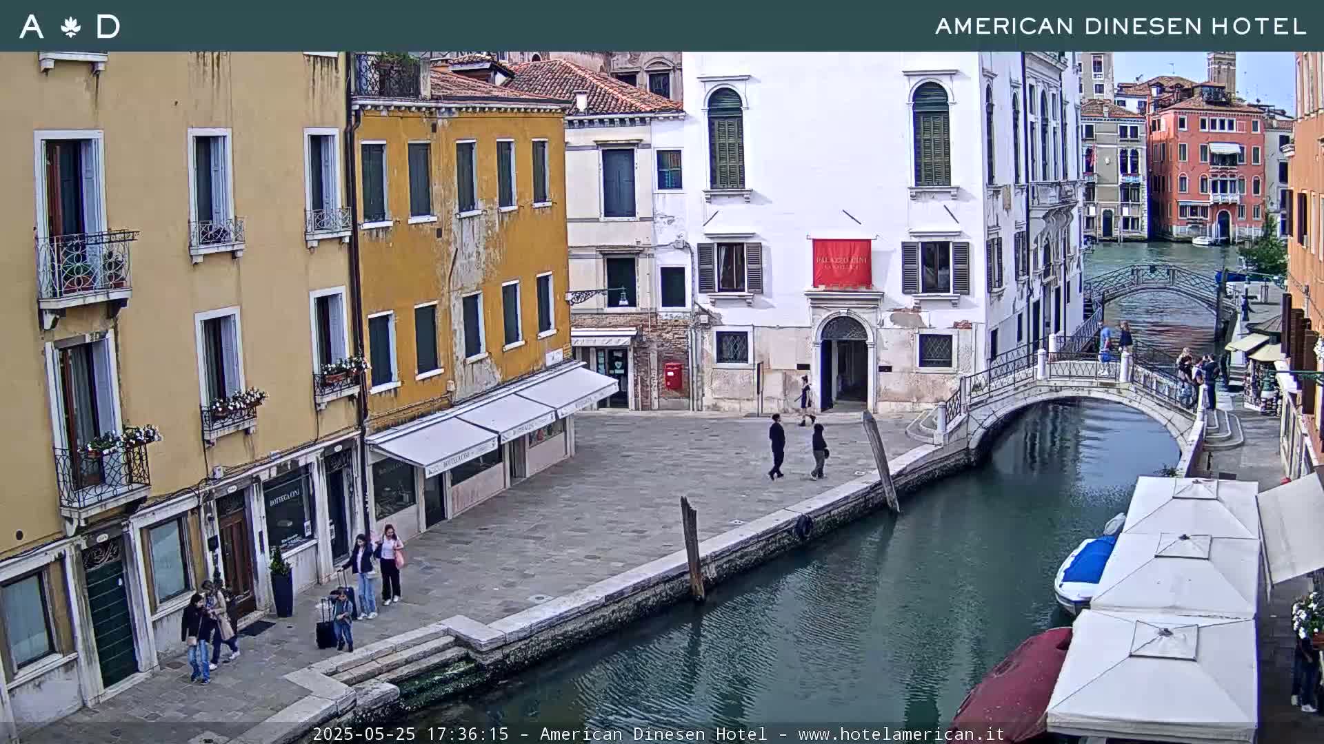 A sunny Venetian canal scene shows several pedestrians near colorful buildings and a small bridge, with boats and outdoor cafe umbrellas visible.