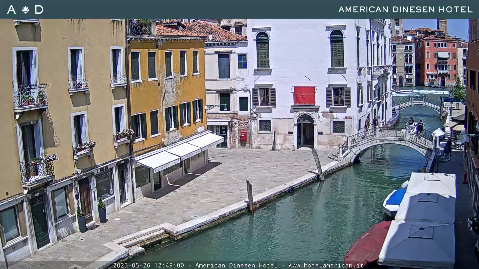 A sunny view of a canal in Venice, Italy, shows buildings lining the waterway and a pedestrian bridge crossing it; boats are moored along the canal bank.