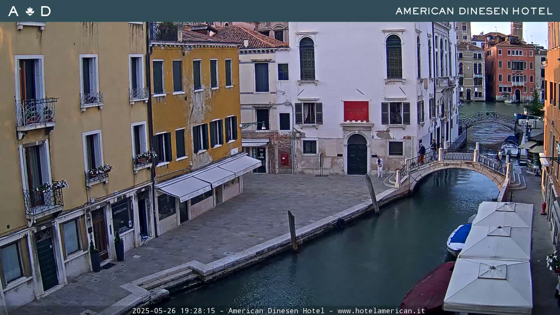 A canal in Venice, Italy, shows several buildings lining the waterway and a small bridge connecting two sides of the canal under a partly cloudy evening sky.