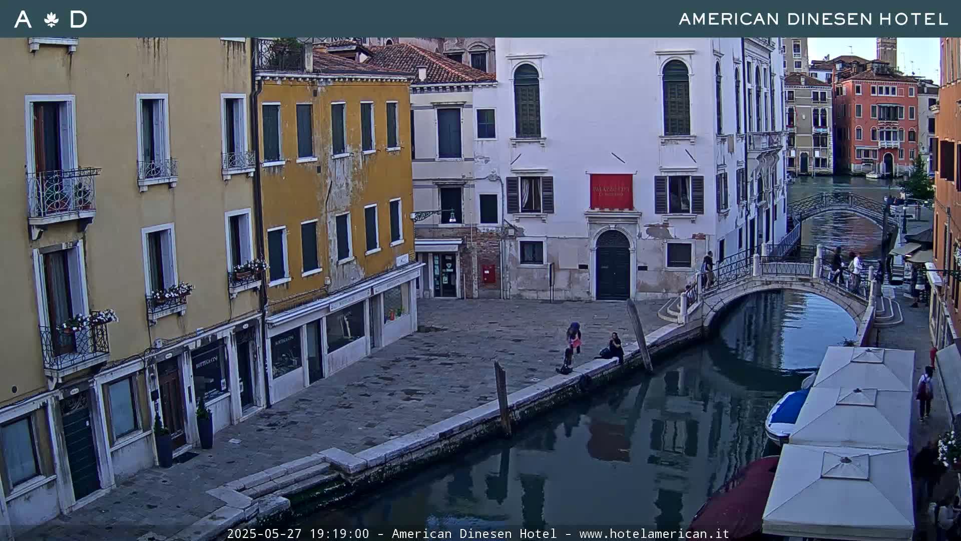 A view of a canal in Venice, Italy, at dusk, showing buildings lining the waterway and a small bridge with people walking on it.