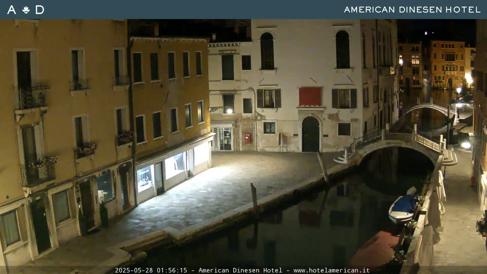 A nighttime view of a canal in Venice, Italy, showing several buildings lining the waterway and a small boat moored alongside a bridge.