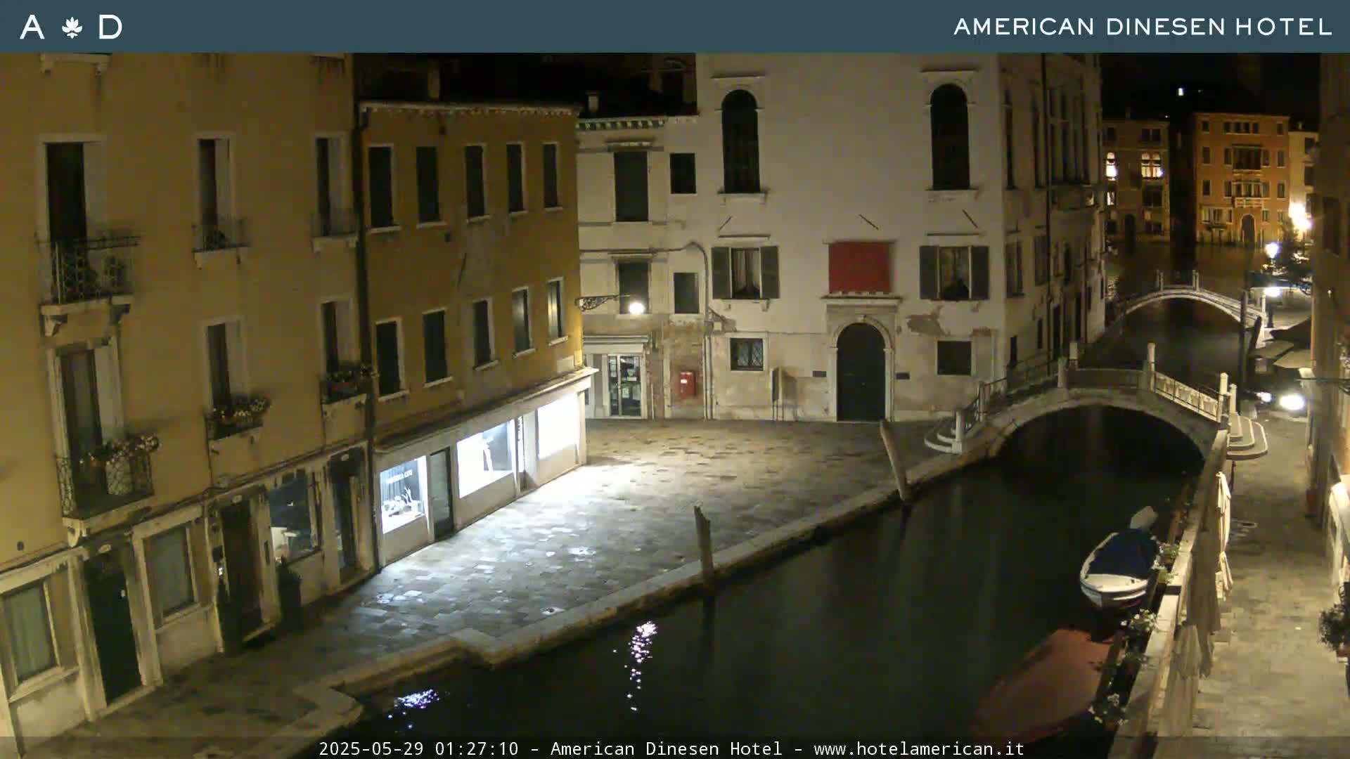 A nighttime view of a canal in Venice, Italy, shows several buildings lining the waterway, with a small bridge and a boat visible in the water.