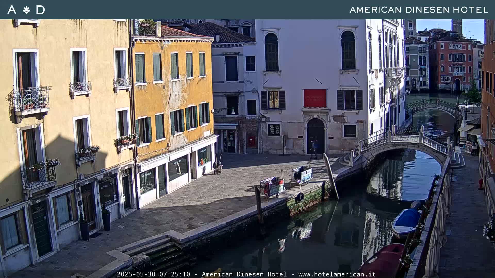 A sunny view of a canal in Venice, Italy, shows several buildings lining the waterway, with a small bridge spanning the canal.