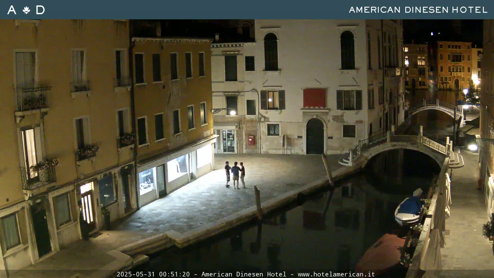 At night, three people stand in a plaza beside a canal in Venice, Italy, which is spanned by a small bridge.