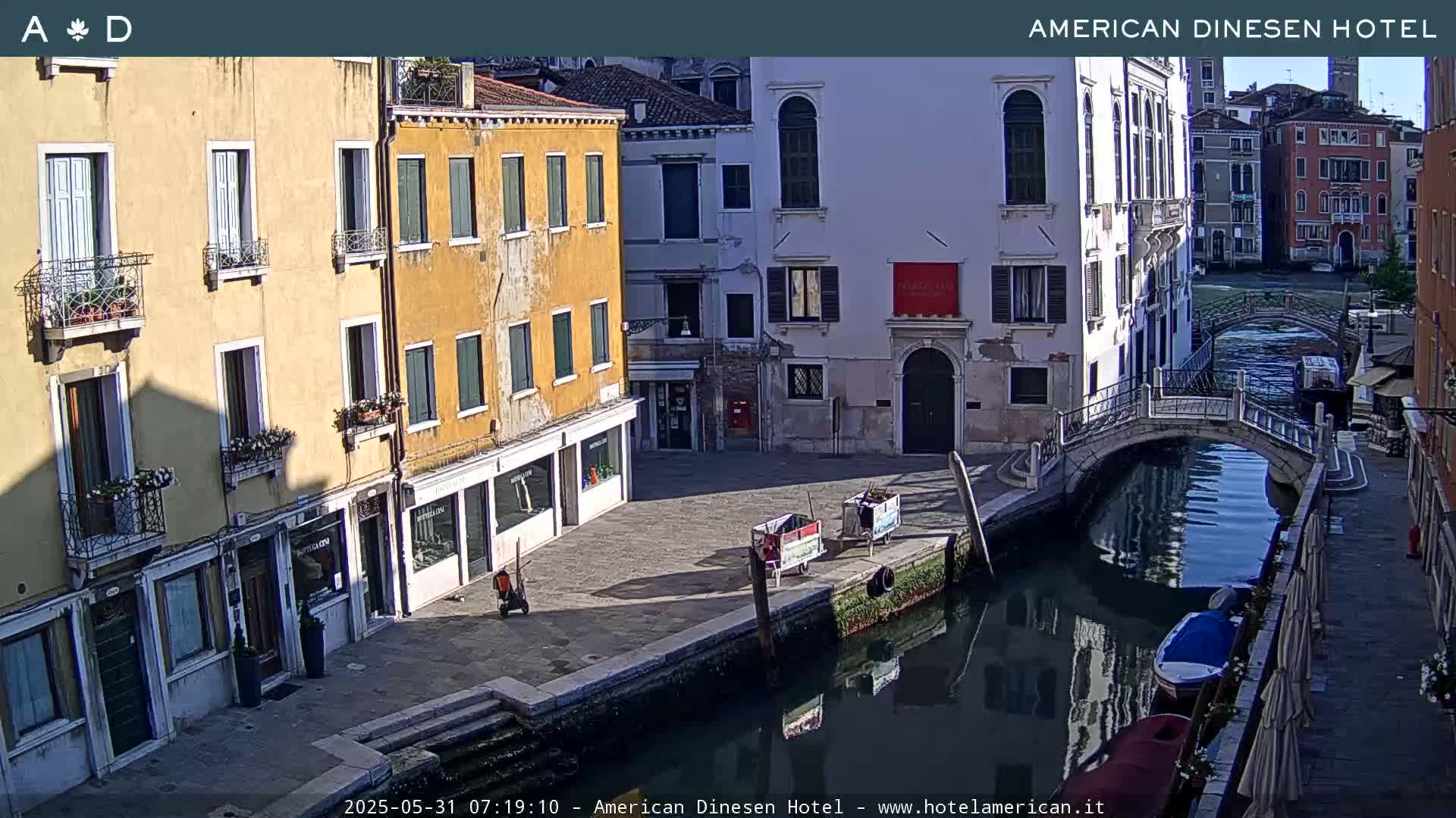 A sunny day in Venice shows a canal with a small bridge, several buildings lining the waterway, and a few small boats.