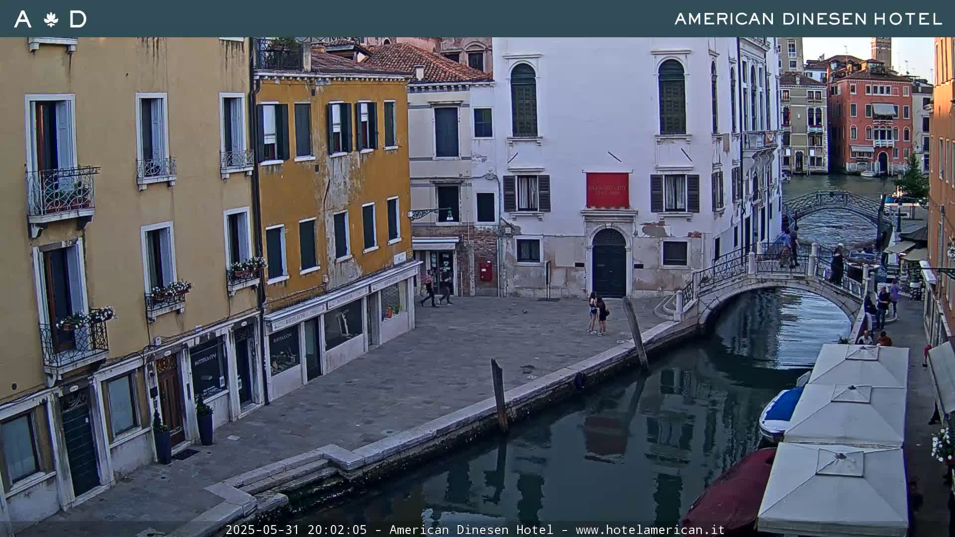 A Venetian canal at dusk, with several buildings lining its banks, a small bridge crossing the water, and a few people walking along the canal-side pavement.