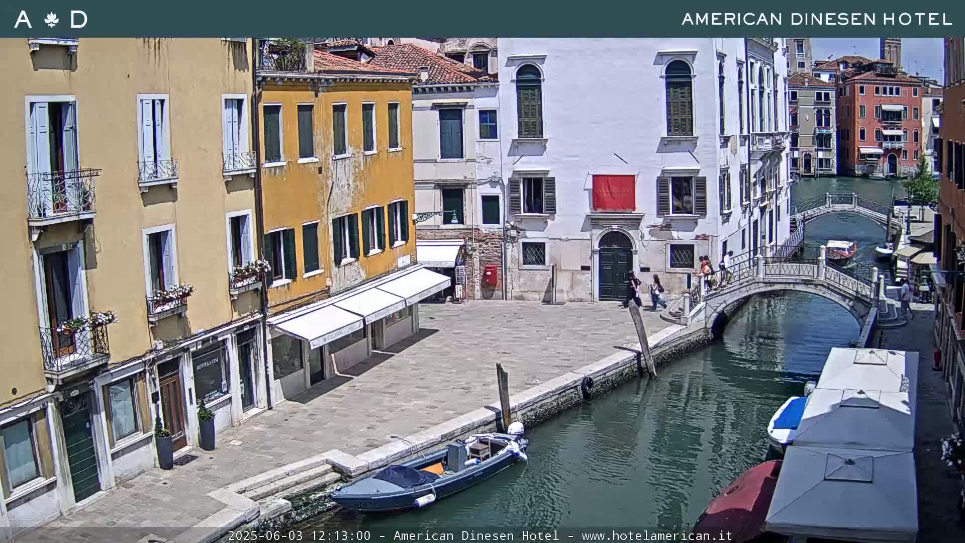 A sunny Venetian canal scene shows several buildings lining the waterway, a small bridge, and a few boats.