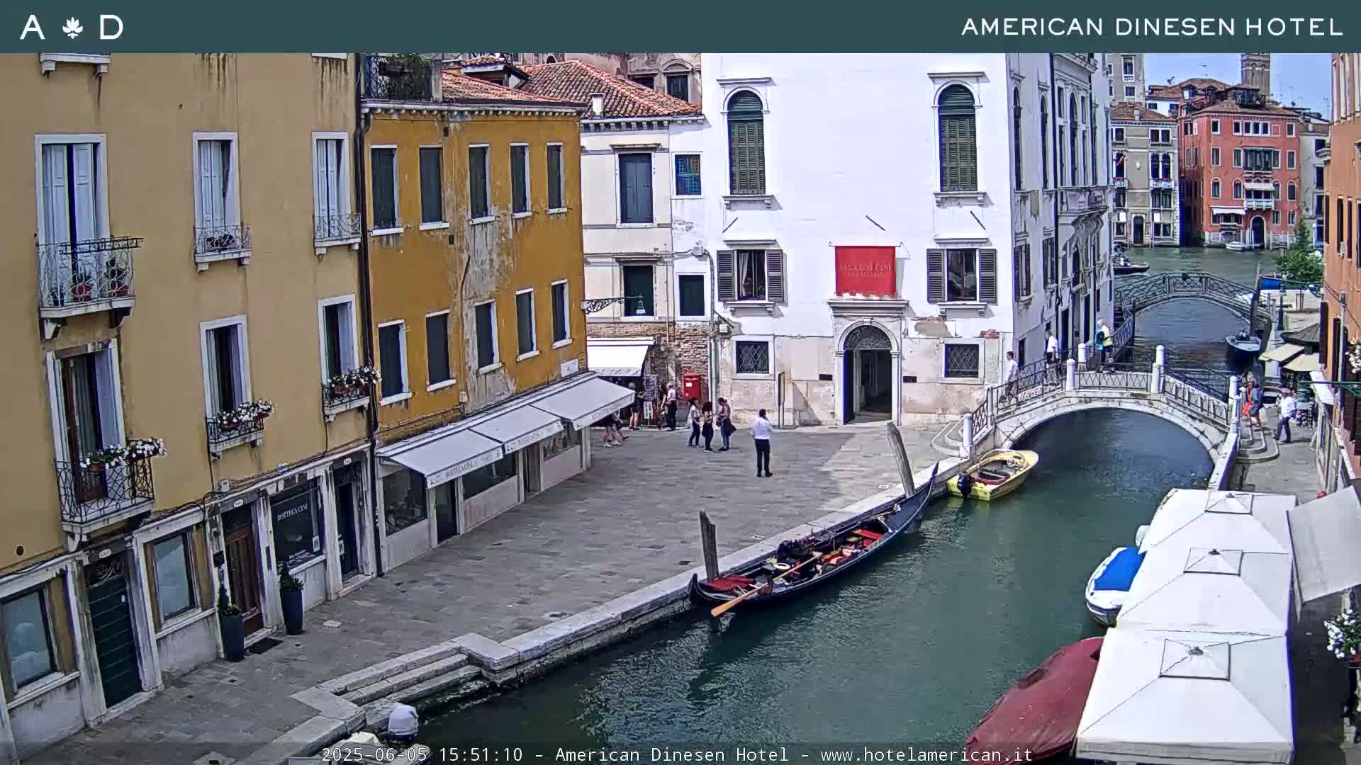 A sunny Venetian canal scene shows a gondola moored alongside a stone embankment, several buildings lining the waterway, a small footbridge crossing the canal, and a few pedestrians.