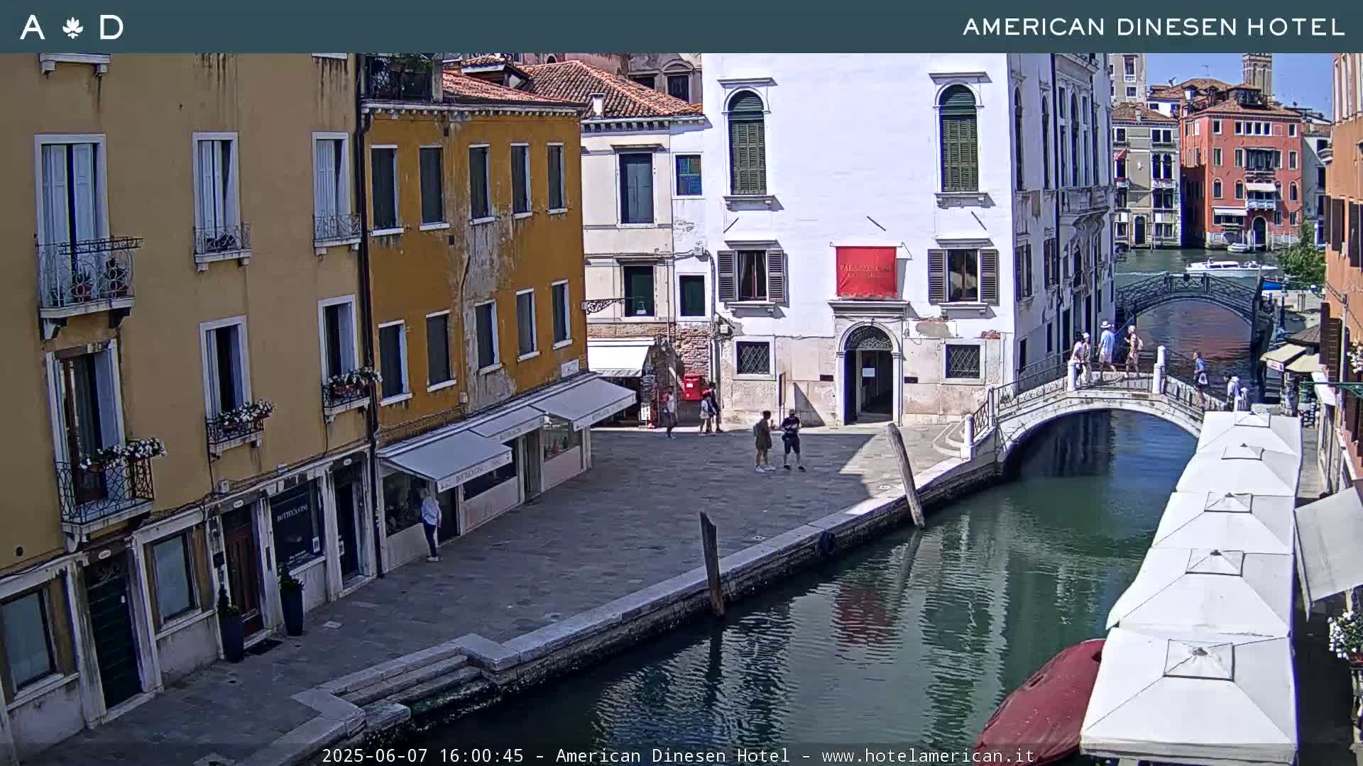 A sunny view of a canal in Venice, Italy, shows several buildings, a footbridge with pedestrians, and outdoor seating areas.