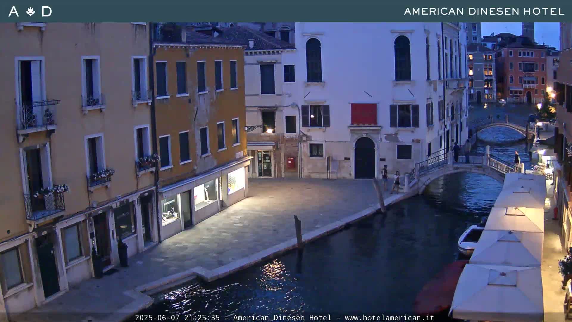 A tranquil canal scene in Venice at twilight, showing several buildings lining the waterway, with a few pedestrians visible on a bridge and along the canal's edge.