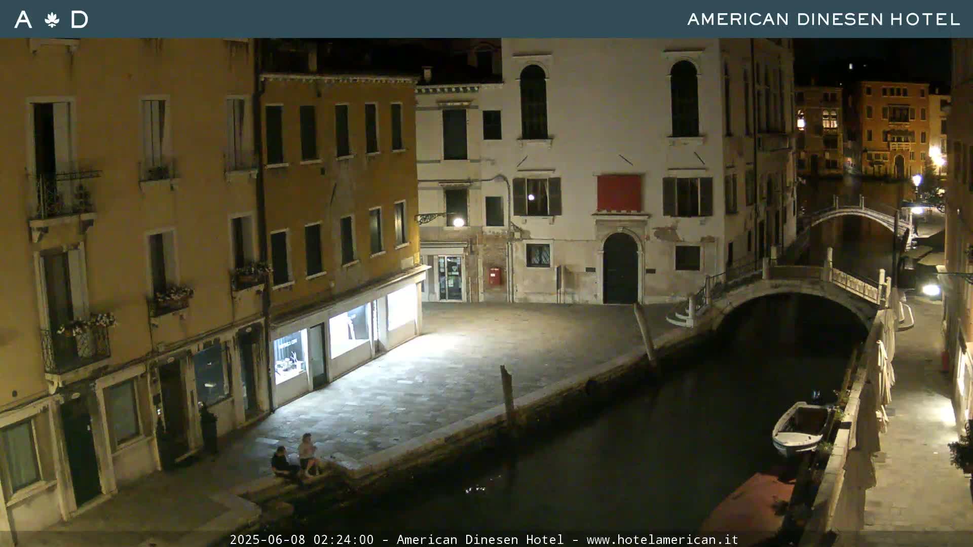 At night, a canal scene in Venice shows two people sitting on a wall by the water, a small boat moored nearby, and several buildings lining the canal.