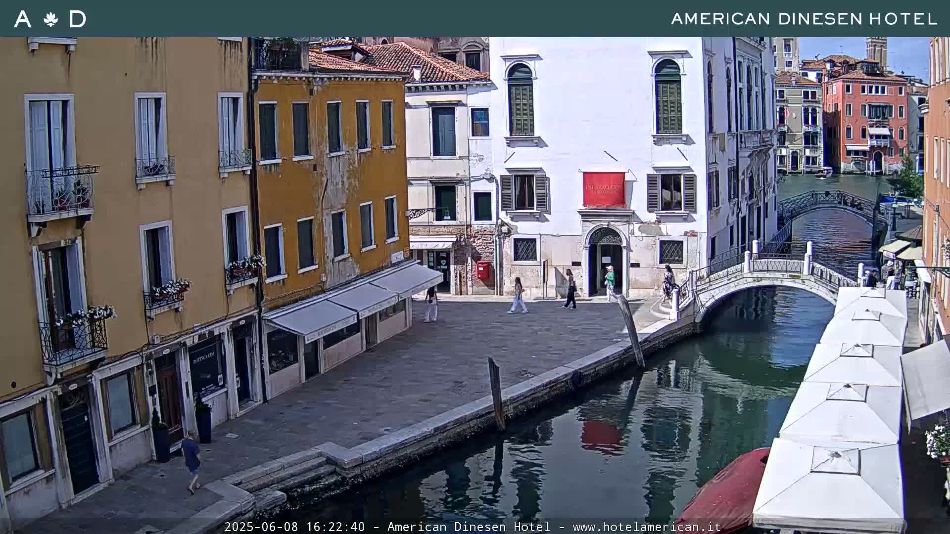 A sunny view of a canal in Venice, Italy, shows several buildings lining the waterway, a small bridge crossing the canal, and a few people walking along the waterfront.