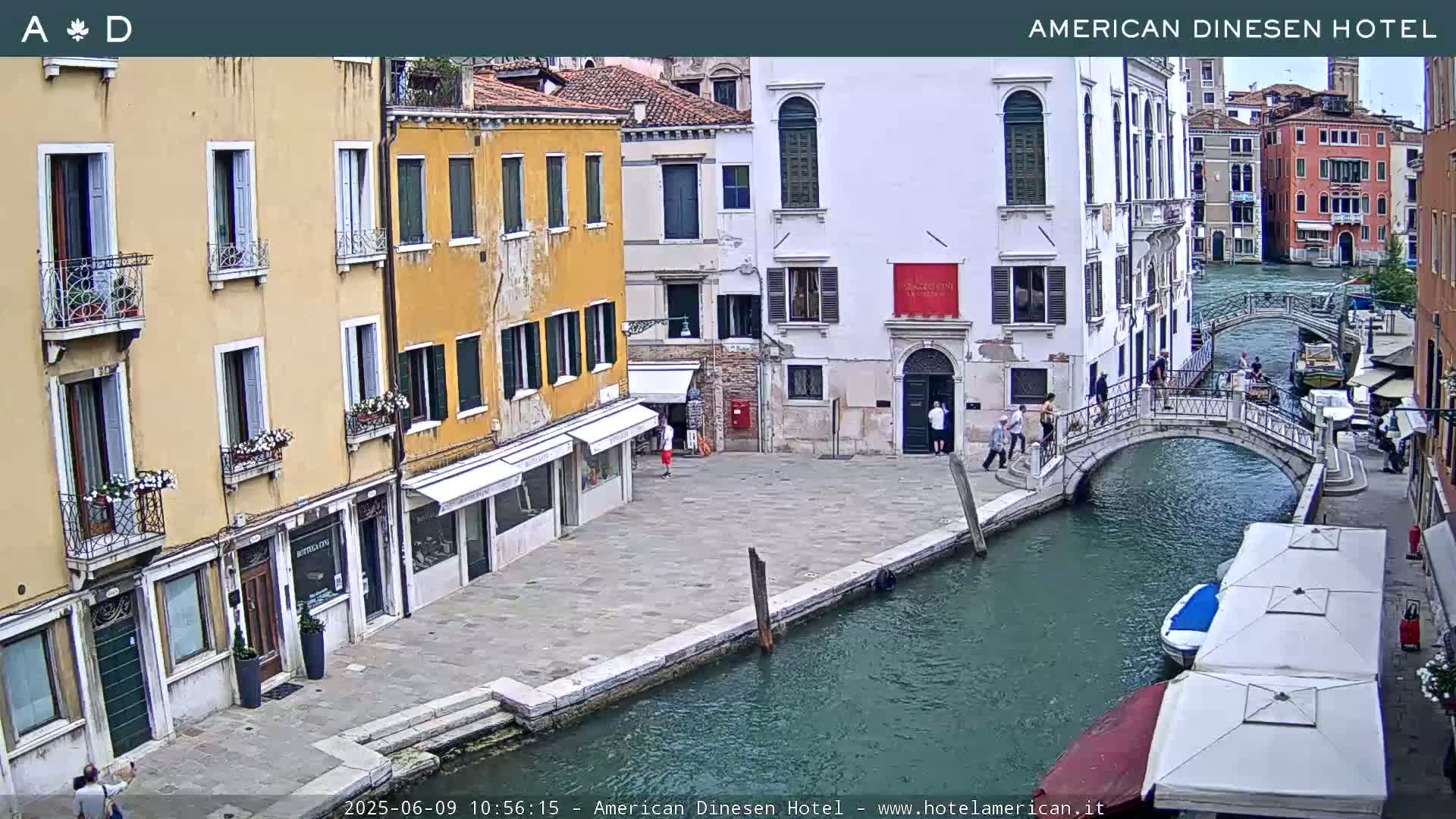 A canal in Venice, Italy, with several buildings lining its banks, a small footbridge spanning the waterway, and boats moored alongside, on a partly sunny day.