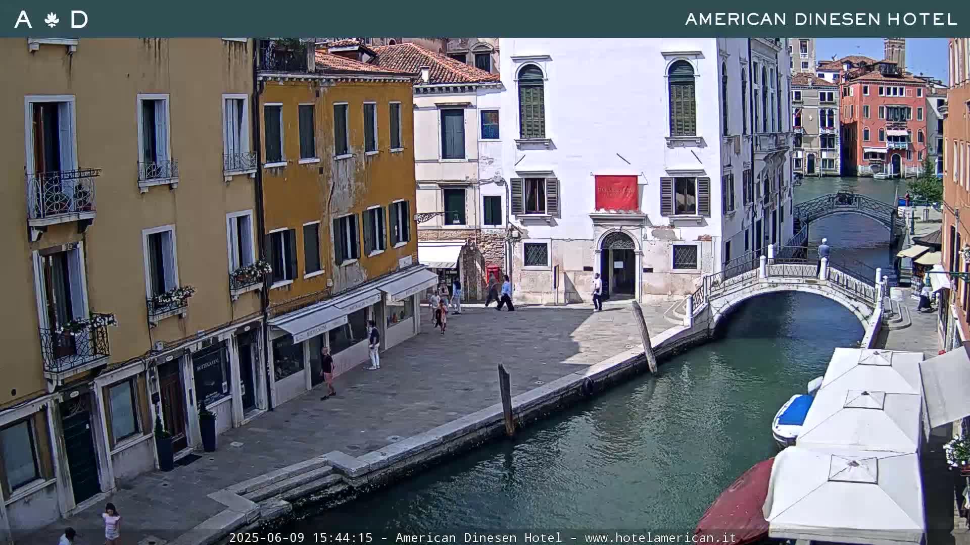 A sunny day in Venice shows several buildings lining a canal, with a small footbridge crossing the waterway, and a few people walking along the canal side.