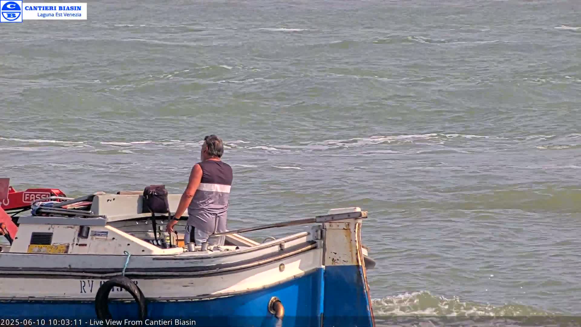 A man stands on the back of a small, white and blue boat on a choppy body of water.