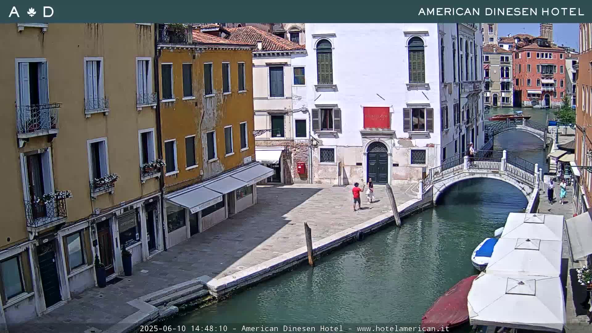 A sunny Venetian canal scene shows colorful buildings lining the waterway, a small footbridge, and a few people walking along the canal's edge.