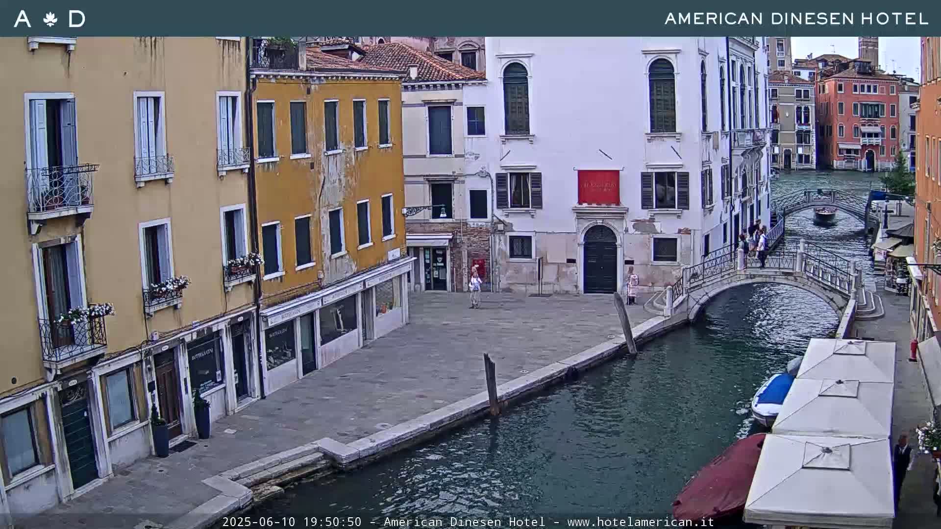 A canal in Venice, Italy, shows several buildings lining its banks, a small footbridge crossing the waterway, and a few boats moored nearby, under a mostly clear sky.