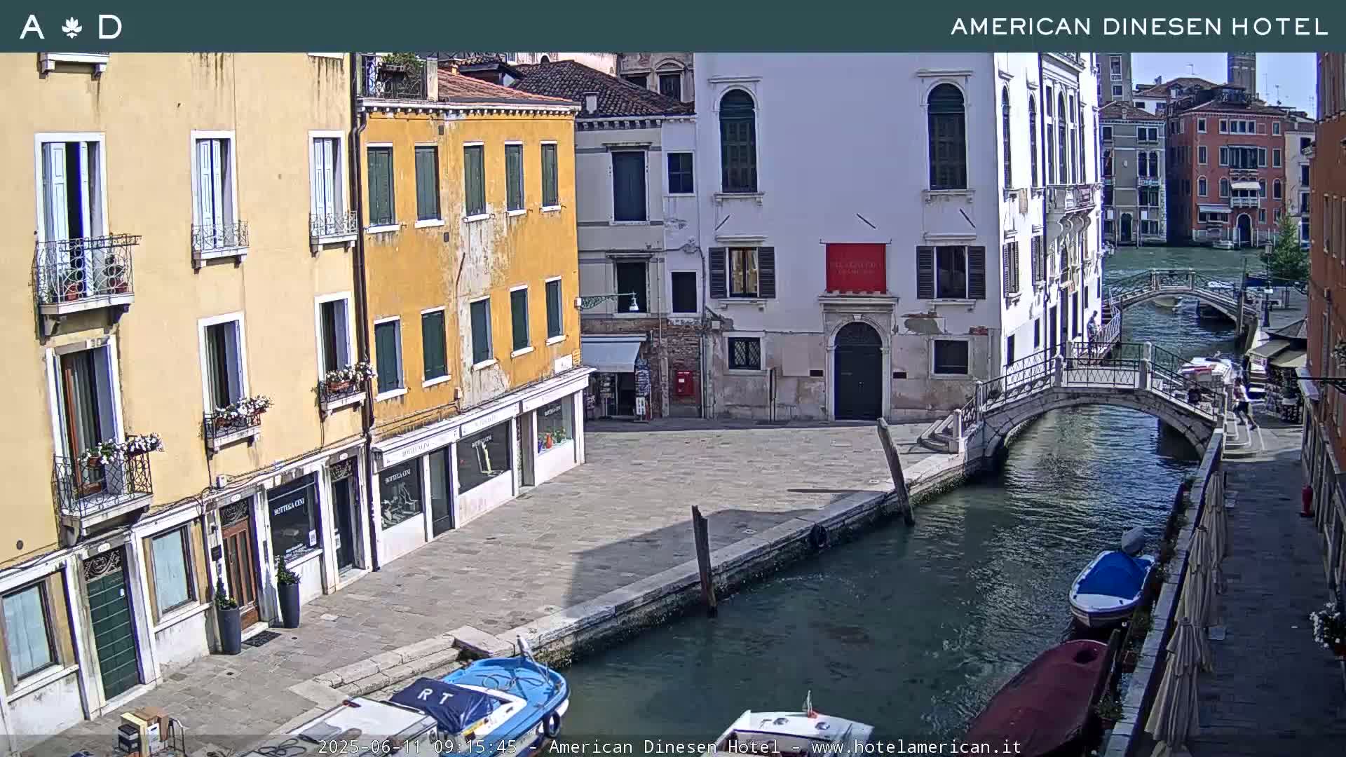 A sunny day in Venice shows a canal with several boats and bridges between buildings with flower boxes on their windowsills.