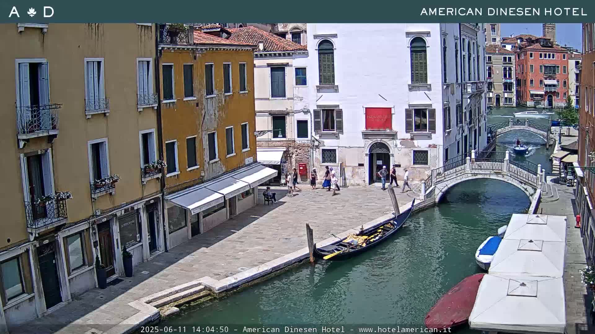 A sunny Venetian canal scene shows a gondola in the water, several people walking along the canal-side, and buildings lining the waterway, with a small bridge crossing the canal.