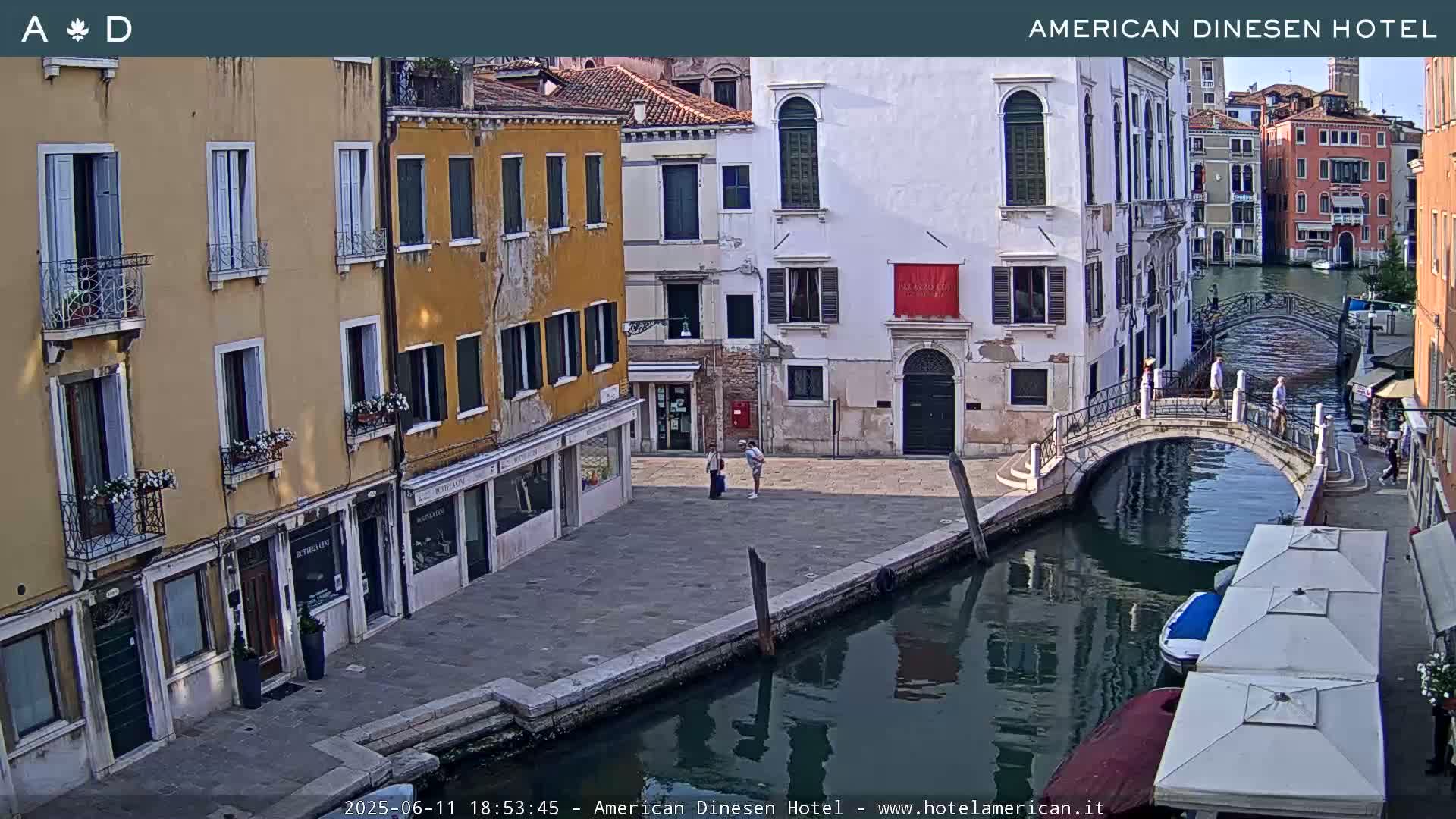 A sunny view of a canal in Venice, Italy, shows colorful buildings lining the waterway, a small bridge, and a few pedestrians.