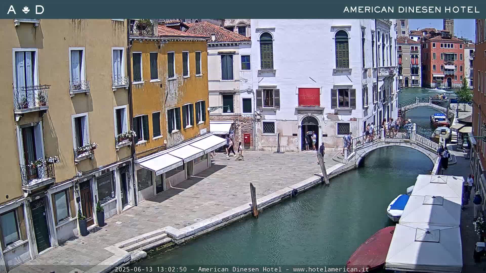 A sunny Venetian canal scene shows several buildings, a footbridge with people on it, and several boats moored along the canal's edge.