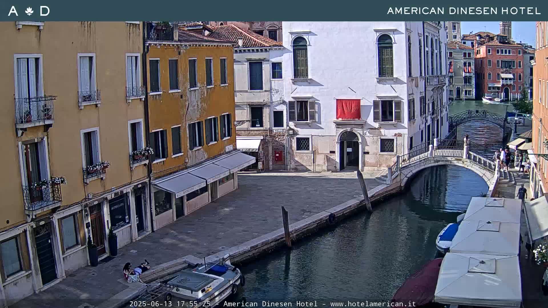 A sunny day in Venice shows a canal with a small bridge, several buildings lining the waterway, and people walking along the canal.