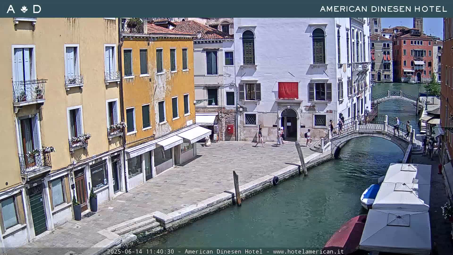 A sunny Venetian canal scene shows several buildings lining the waterway, a pedestrian bridge, and a few people walking along the canal.