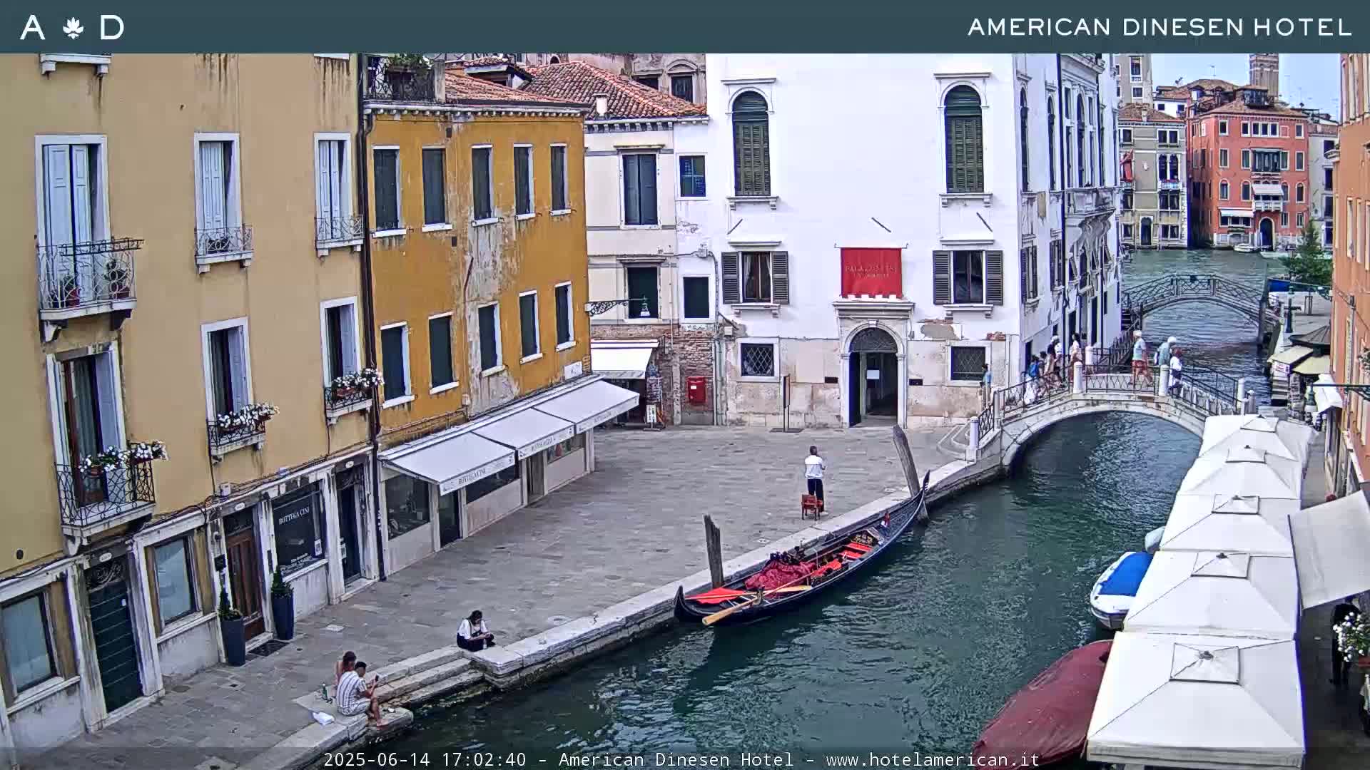 A gondola is moored along a canal in Venice, Italy, under a partly cloudy sky, with several buildings lining the waterway and people strolling on a nearby bridge.