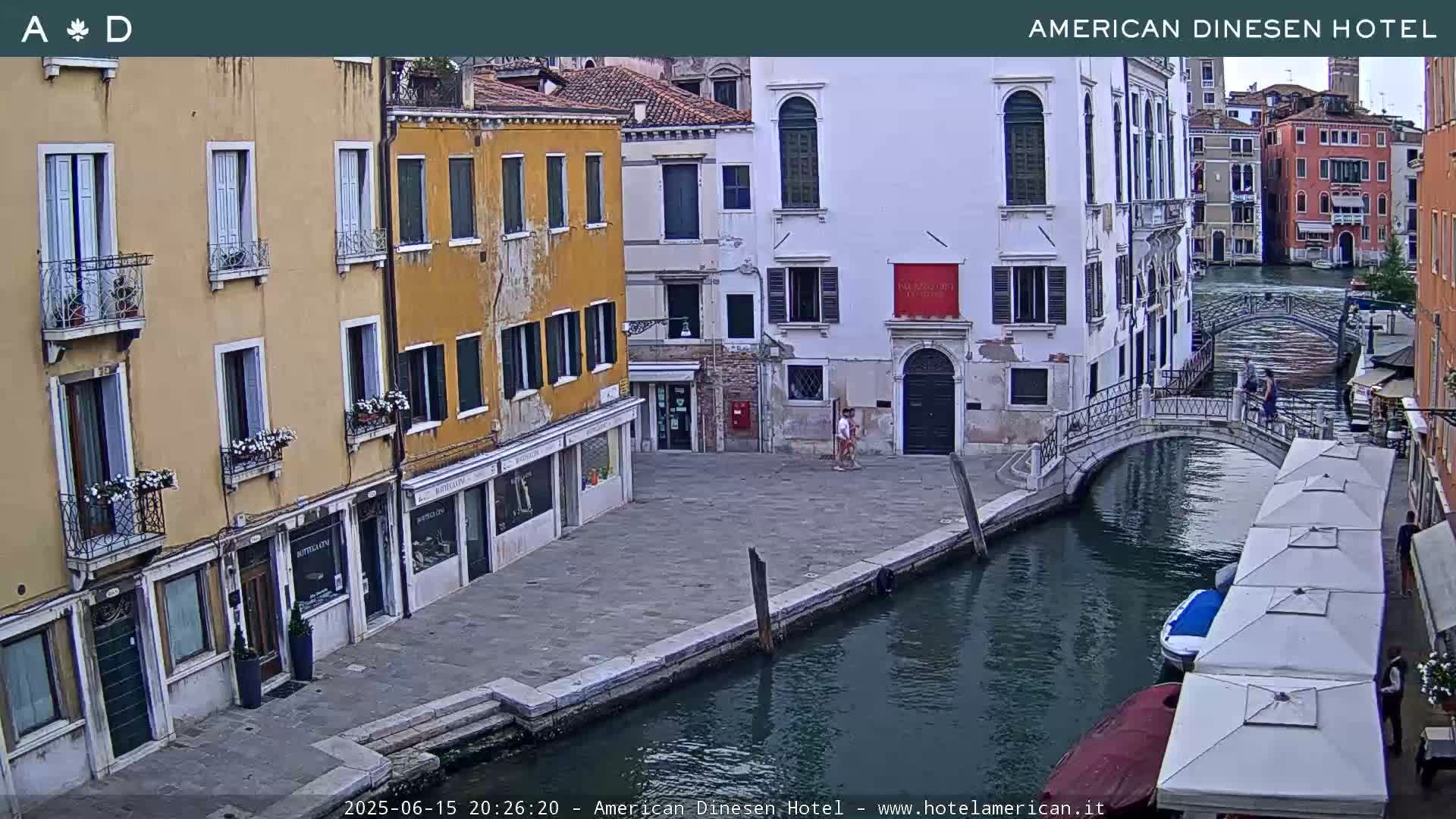 A canal in Venice, Italy, under a twilight sky, is bordered by buildings with several people visible on a footbridge and along the canal.