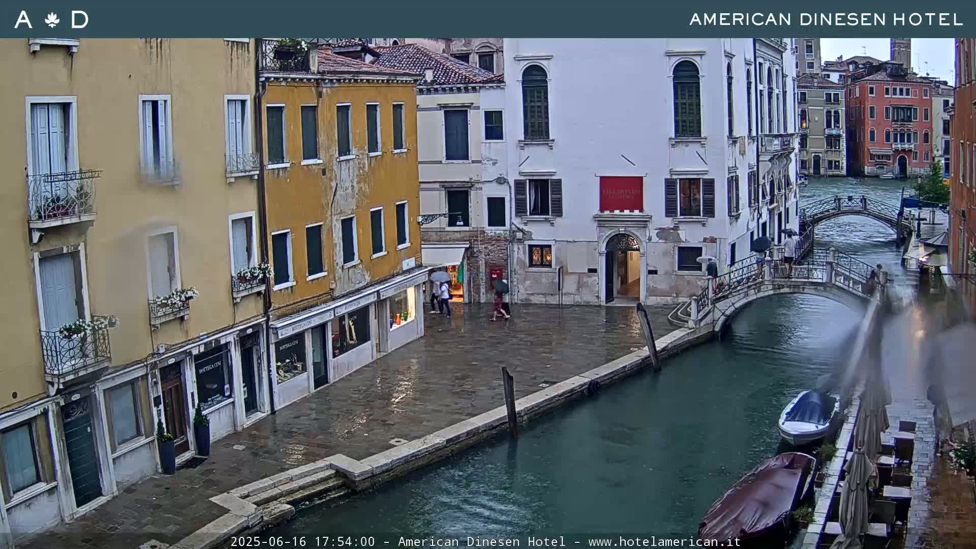 A rain-slicked Venetian canal scene shows several buildings lining the waterway, pedestrians with umbrellas, and a small boat moored along the canal bank.