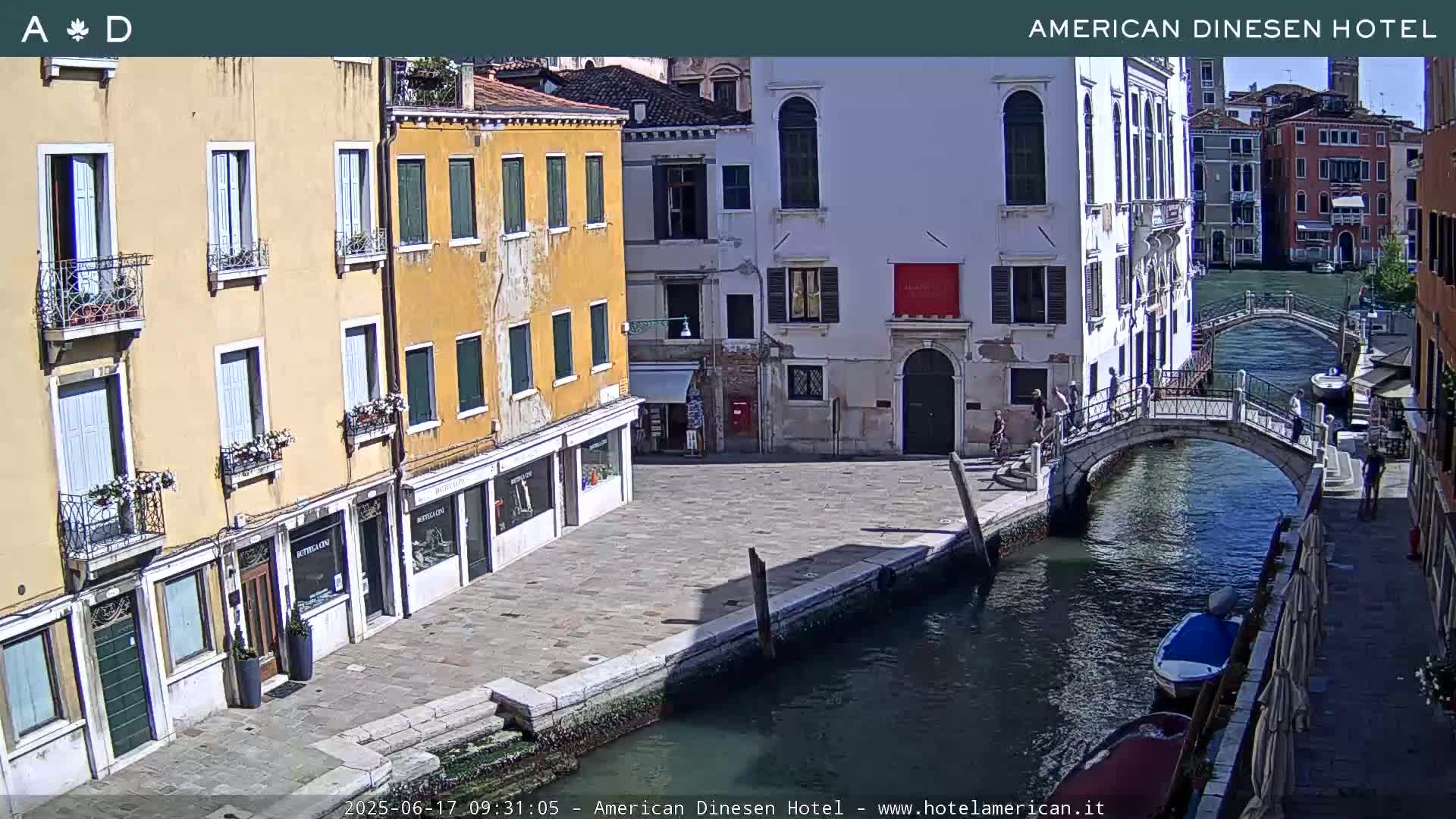 A sunny Venetian canal scene shows several buildings lining its banks, a footbridge spanning the water, and small boats moored along the waterway.