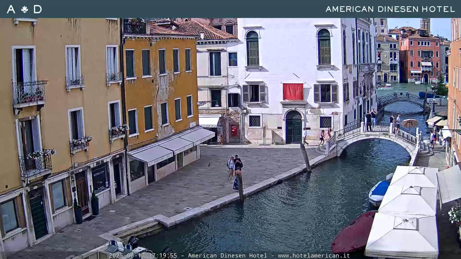 A sunny day in Venice shows a canal with a small bridge, pedestrians, and buildings lining the waterway.