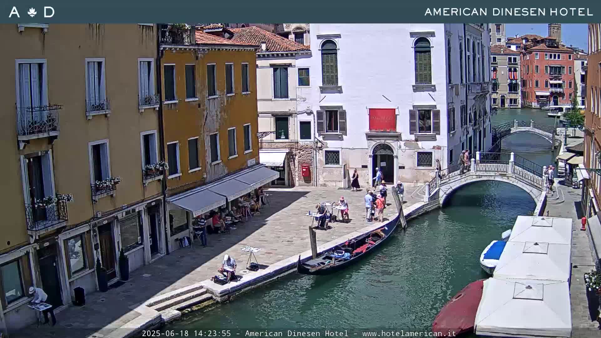 A sunny day in Venice shows a canal with a gondola and several people on a stone walkway next to colorful buildings and a small bridge.